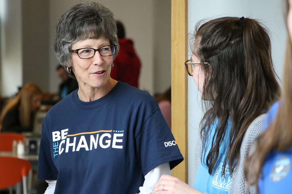 Melanie Sipko talks to a student. Sipko has short salt and pepper hair and wears a t-shirt that reads "Be the Change."