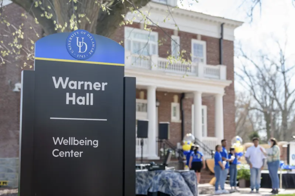 The Wellbeing Center at Warner Hall sign in the foreground. The brick building with white trim is in the background. 