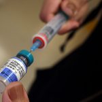 A syringe pulls liquid from a bottle containing vaccine