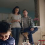 A young child with autism stands in the foreground with its parents watching from behind