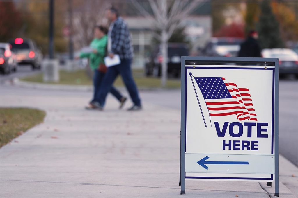 Vote here sign in foreground with people in the background walking in direction sign is pointing towards