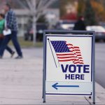 Vote here sign in foreground with people in the background walking in direction sign is pointing towards