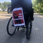 A person using a wheelchair displays a sign reading, We Rise Together.