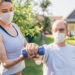 A home care aide wearing a mask assists an elderly man wearing a mask doing an arm exercise.