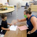 A teacher sits across a desk from a student with a clear plexiglass divider between them