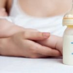 A woman cradles an infant in her arms. In front of them sits a bottle of milk.