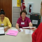 Two poll workers sit at a table with papers scattered in front of them