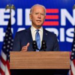 President-elect Joe Biden delivers a speech after being named the winner of the 2020 election. Biden stands in front of a large version of his campaign logo and an American flag. He speaks from a podium with a thoughtful expression.