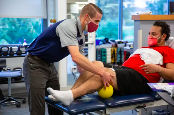 In a physical therapy clinic, a college student wearing a mask bends over and touches the knee of an older man sitting facing him.