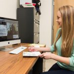 A young woman sits at a desk taking notes as she speaks to a person via video chat