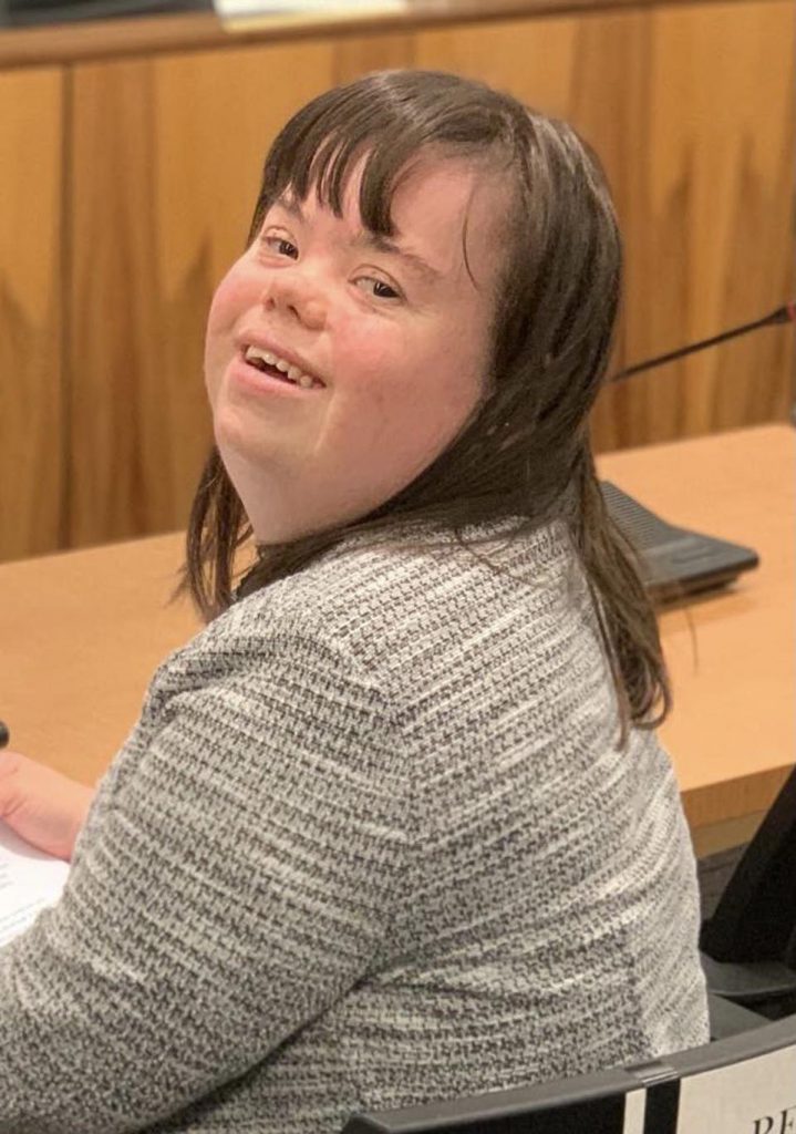 Charlotte Woodward, a disability advocate with Down syndrome, looks over her shoulder smiling at the viewer