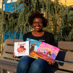 A Black woman sits on a bench with picture books spread out on her lap. Each book cover has a nonwhite character pictured.