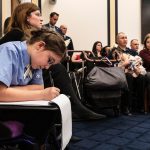 A young woman in a wheelchair writes on a notepad. She is in a busy waiting room.