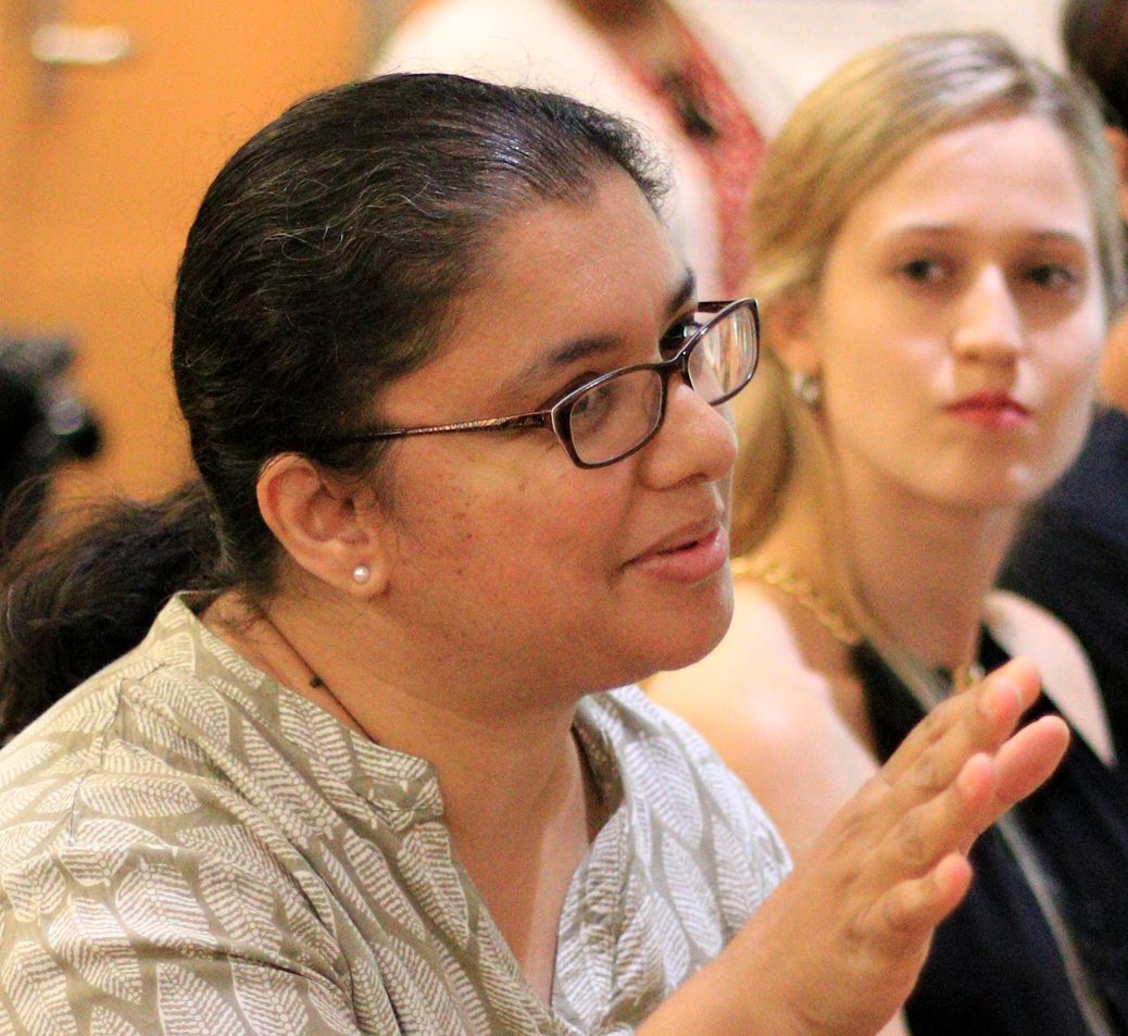 A professor gestures from her seat as she speaks to a group sitting around a large table. A young blonde woman looks on from the background