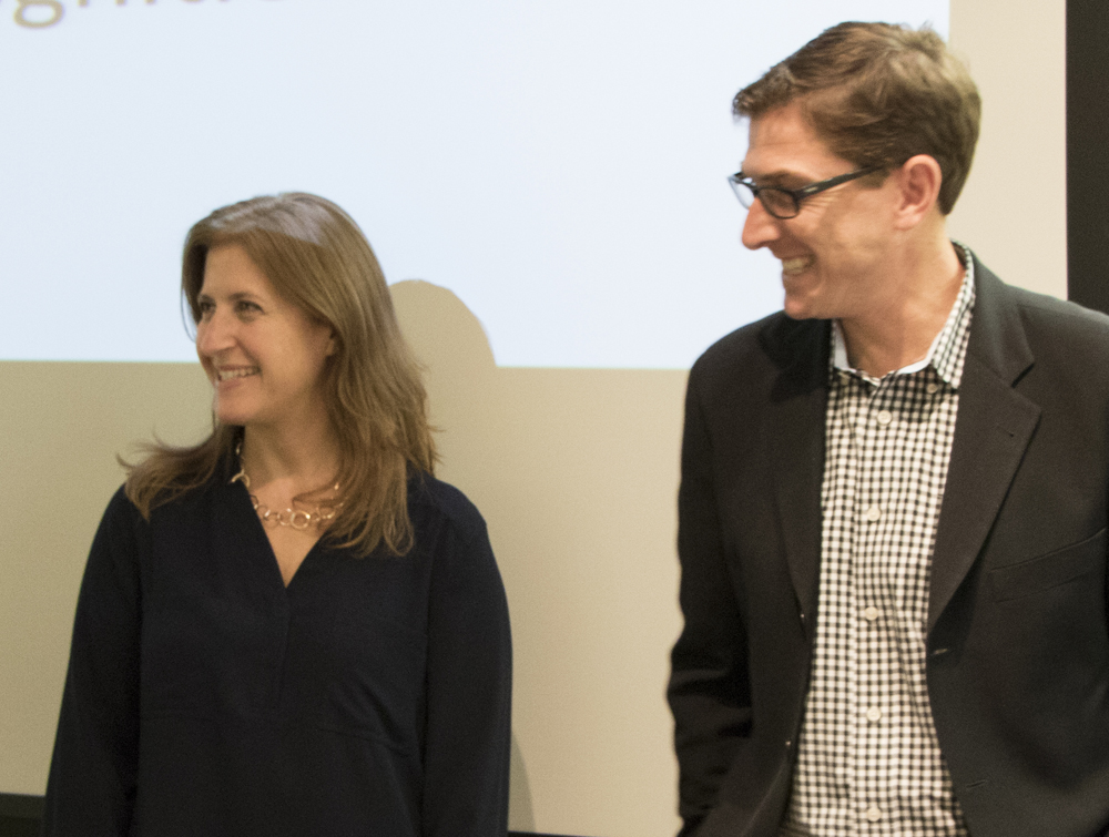 Two professors, a woman and a man, stand together in front of an overhead projection. Both smile and look to their right