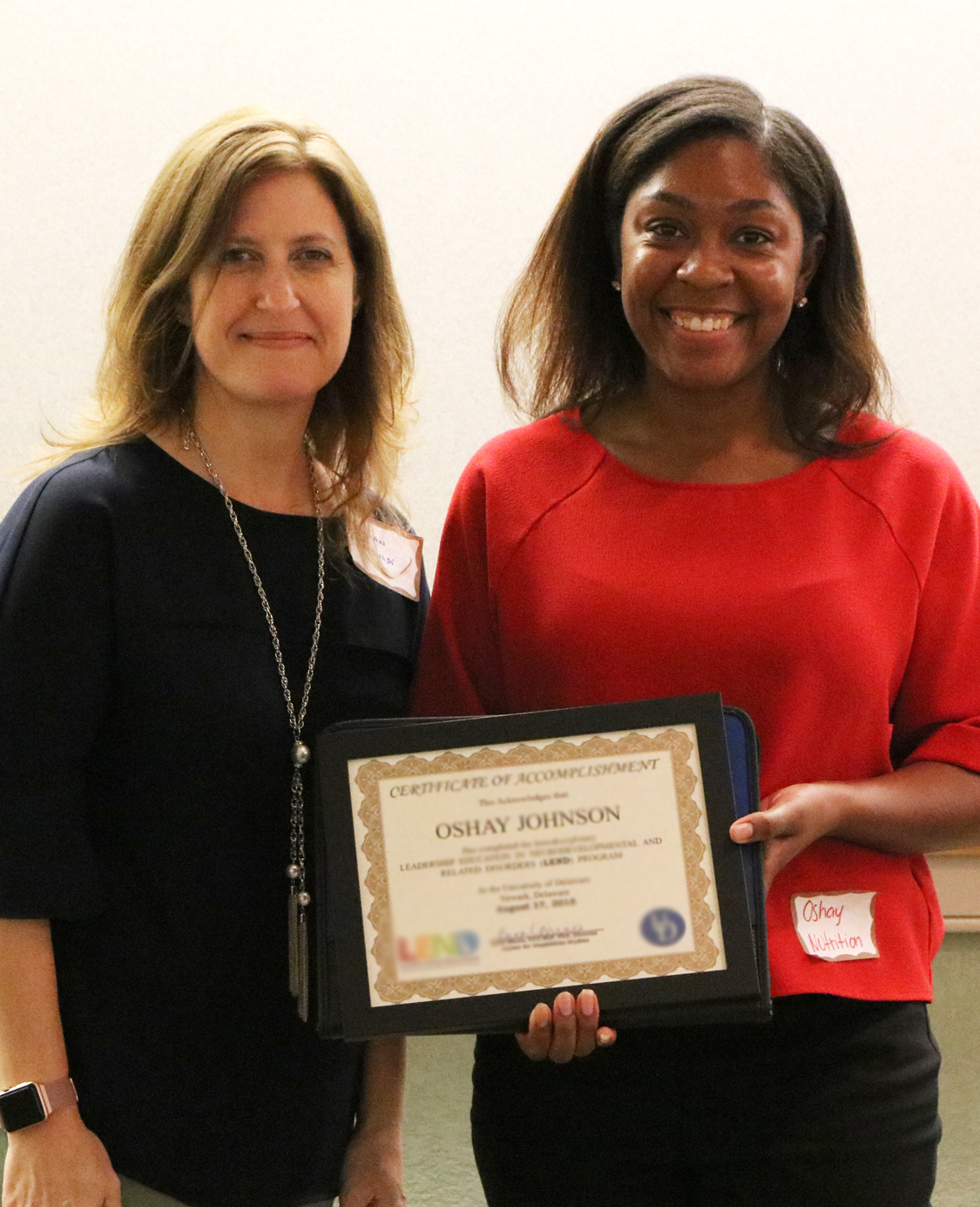 A middle age white woman stands next to a young woman of color, her student. The young woman is holding a certificate. Both smile.