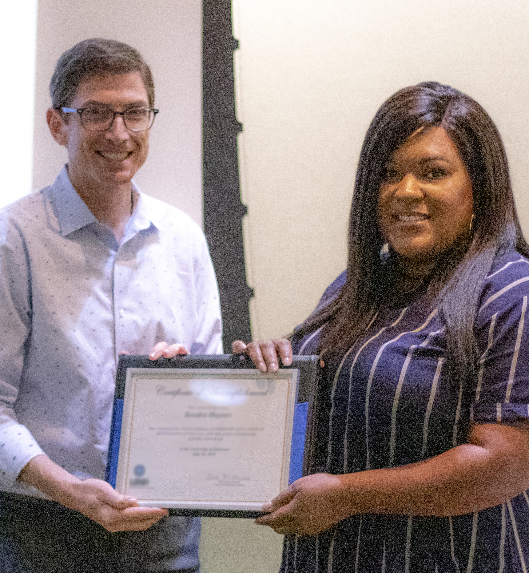 A man in a button down shirt and slacks holds a certificate with a young woman of color.