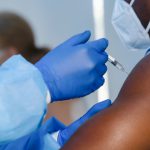 A Black man holds his bare arm out for a medical worker in a sterile gown, goggles and gloves to give him a shot.