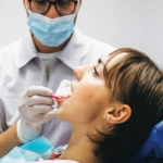 A woman with her head tilted back in a dentist's chair opens her mouth. To her side a dental worker in a white lab coat and face mask sticks a piece of gauze in her mouth.