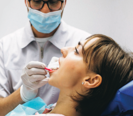A woman with her head tilted back in a dentist's chair opens her mouth. To her side a dental worker in a white lab coat and face mask sticks a piece of gauze in her mouth.