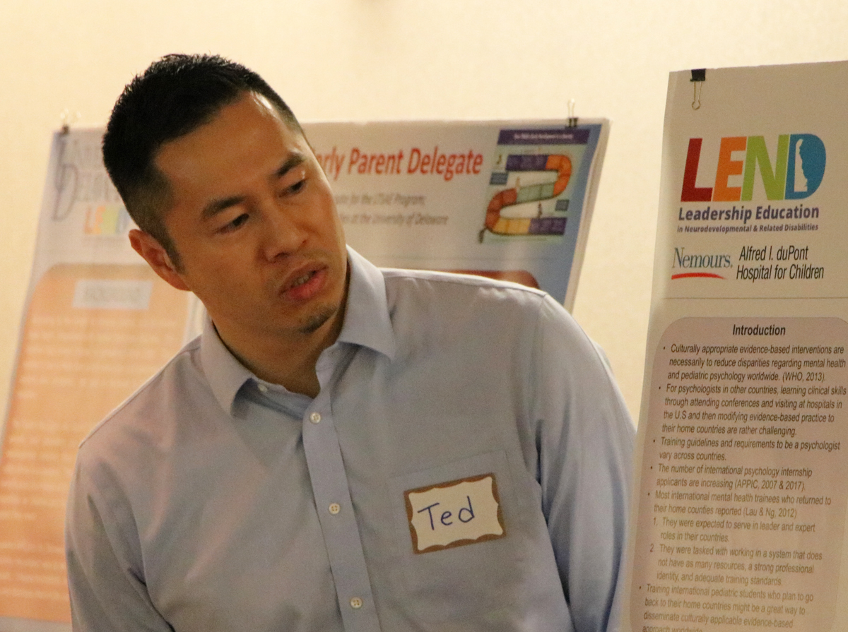 A man of Asian descent in a button down shirt and a nametag reading Ted leans to look at his poster as he gives a research presentation