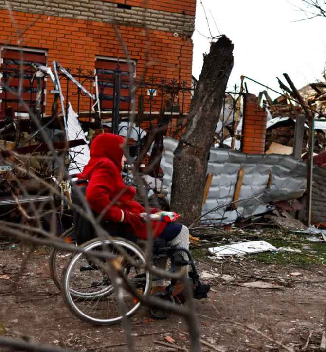 A woman in a red coat sits on her wheelchair beside houses destroyed by Russian shelling amid Russia's invasion of Ukraine, in Sumy