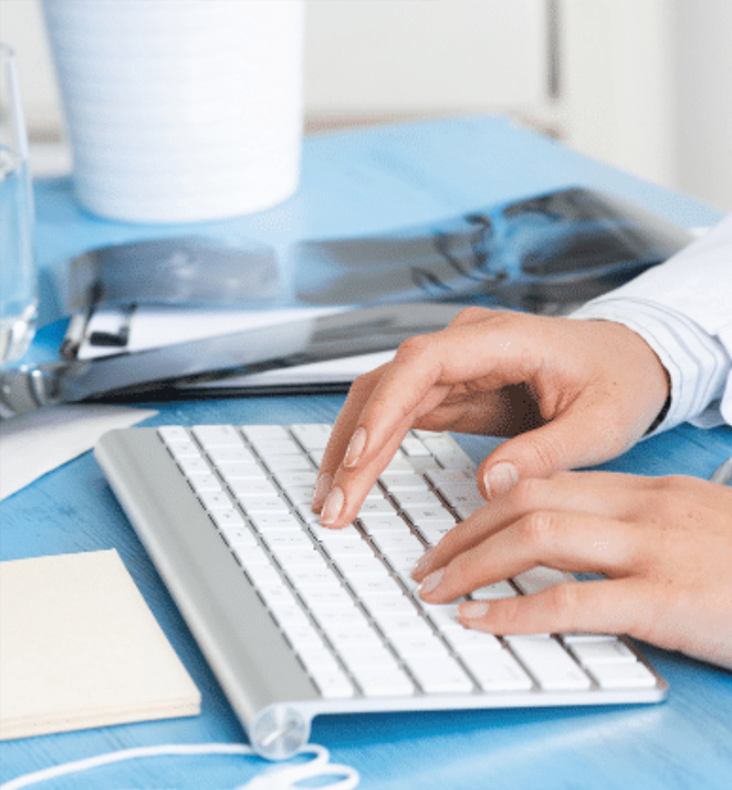 A pair of light-skinned hands type on a keyboard. An x-ray and clipboard are also on the desk.