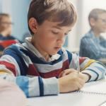 A boy with sandy blond hair in a classroom looks down at his notebook while he holds a pen.
