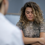 A college-age woman with curly blonde hair wearing a t-shirt with a reptile print crosses her arms and looks down. In the foreground is the blurry suggestion of a woman in a white lab coat.