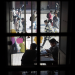 Looking out a window onto two people at a table. Beyond them is a glass wall and people walking in the street or seated on the ground.