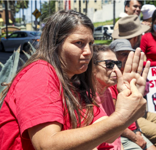 A middle-aged woman in a red t-shirt presses her right index finger into her upraised left palm.