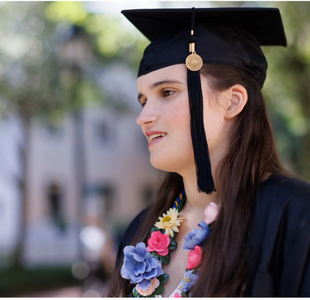 A college-age woman with long dark hair wears a black graduation gown and mortar board and a flower necklace.