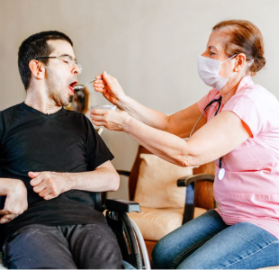 A young adult with short dark hair and some facial hair, seated in a wheelchair, opens his mouth as an older woman in a face mask with a stethoscope feeds him with a spoon.