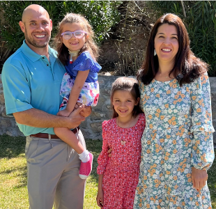 A family of four stand in the sunshine. A little girl with glasses is held by the father on the left. A girl who appears slightly older stands with the mother on the right.