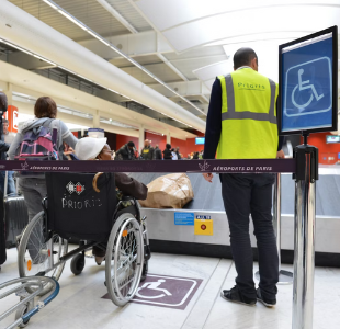 A person in a wheelchair waits with a staff member in a safety vest in the disabled section of the baggage carousel.