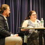 Emily Ladau and Andy Jenks sit on stage in front of a blue velvet curtain. Andy has red hair and wears a blue blazer and is looking at Emily as she speaks. Emily has curly brown hair and wears glasses. She faces the audience.