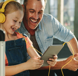 A smiling man in a chambray shirt leans over the shoulder of a girl wearing yellow headphones plugged into a tablet.