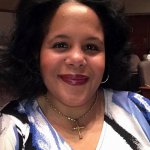 Melissa Gates has dark curly hair and wears red lipstick and a gold cross pendant.