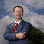Brian Heffernan who has Down syndrome, attended Mass Bay Community College, stands outside of the college. Heffernan wears a blue blazer, white shirt and red tie, and a silver Irish crucifix on a chain around the neck.