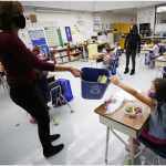 A teacher, almost in silhouette, stands in a classroom. She holds out a recycling bin for a young student with long dark hair, wearing a mask, to drop in a paper.