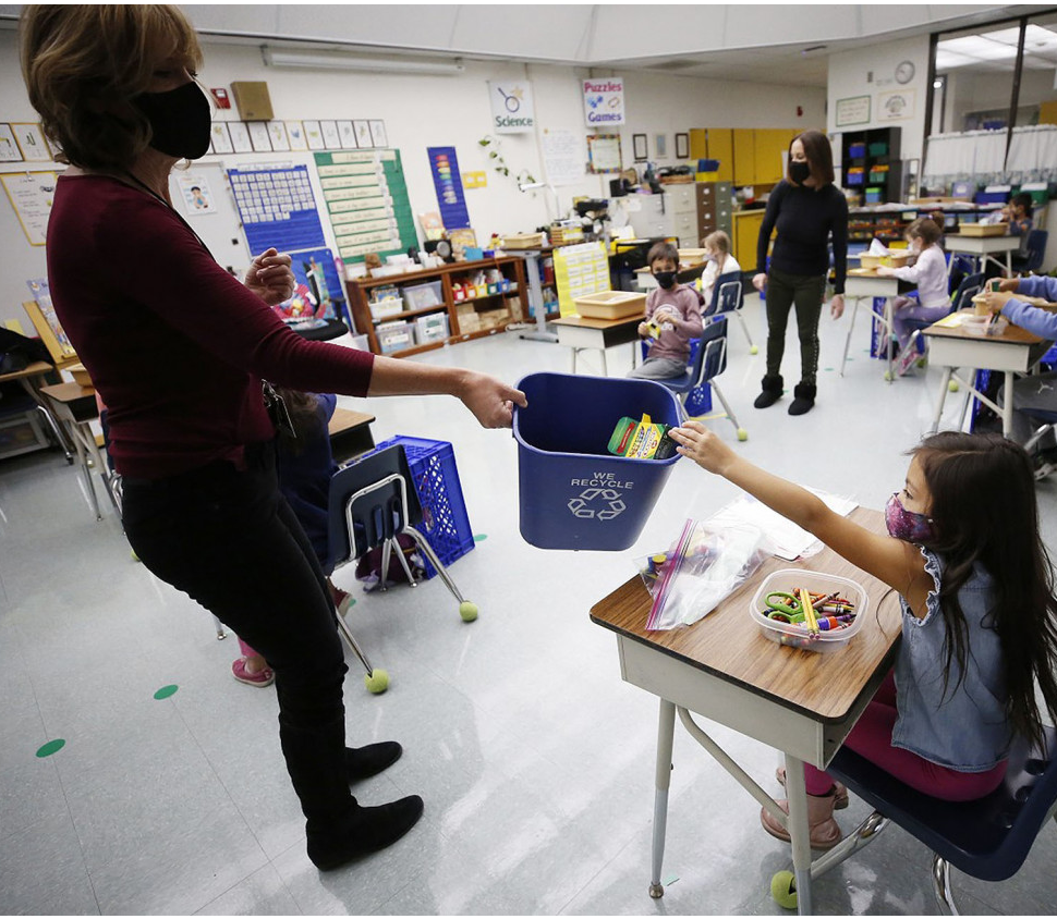 A teacher, almost in silhouette, stands in a classroom. She holds out a recycling bin for a young student with long dark hair, wearing a mask, to drop in a paper.