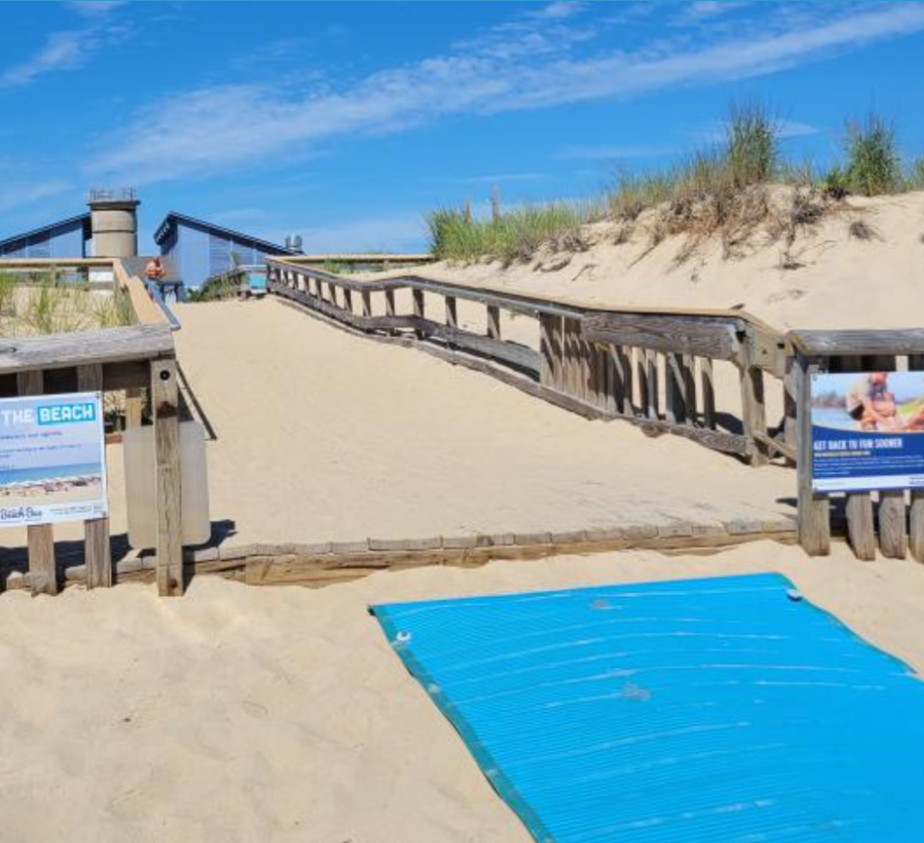 A dune crossing at the Delaware Seashore State Park. Just visible below sand are the wooden boards that make up the ramp. Worn wooden handrails line the ramp.