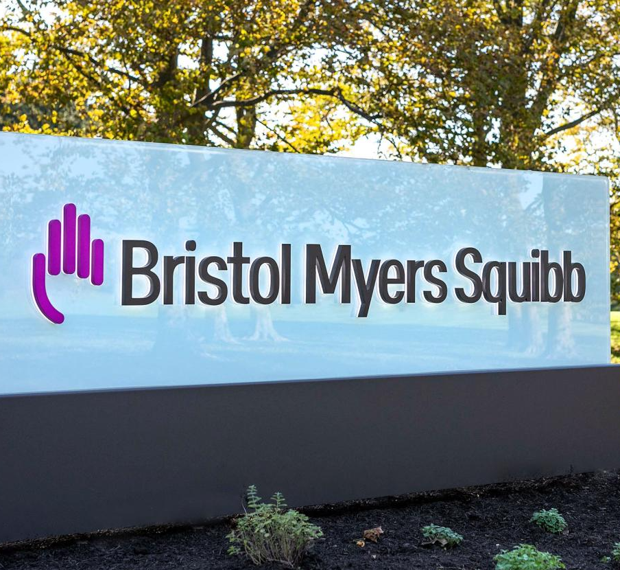 A large exterior sign for Bristol Myers Squibb sits in a landscaped area under large trees.