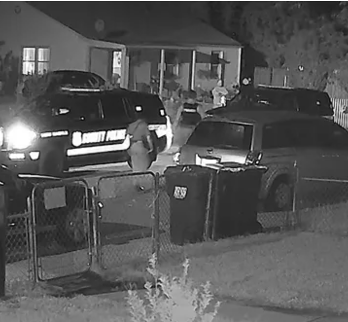 Black and white still from nighttime video footage shows two New Castle County Police SUVs with headlights on in a residential street. The grainy footage looks over a small front yard with chain link fence, trash cans left for pickup and a few parked vehicles. A few police officers and another person are visible.