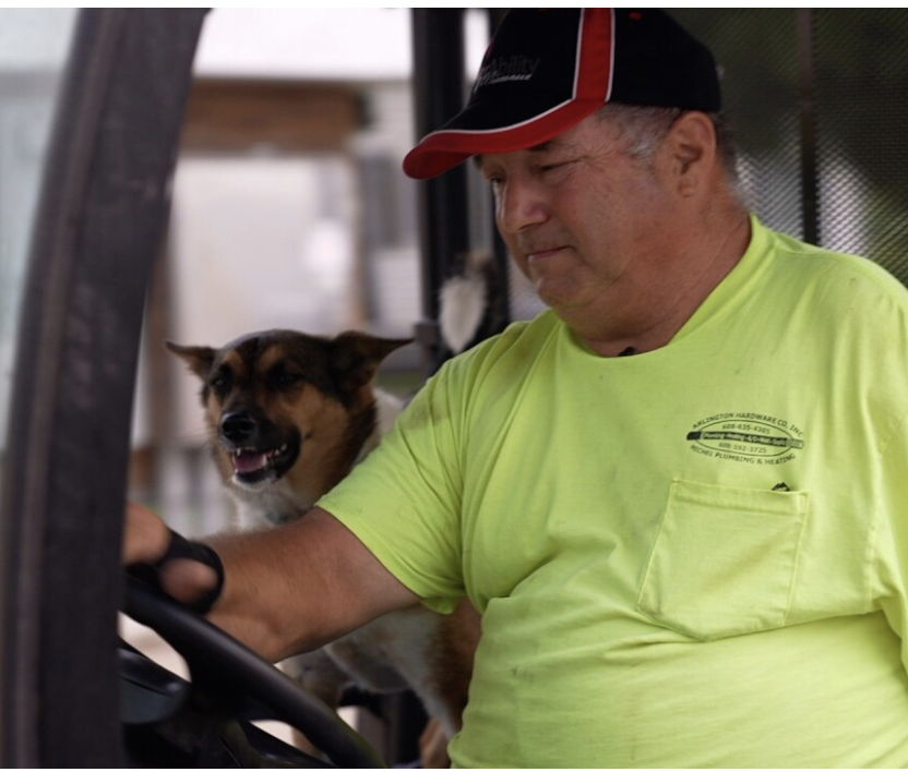 A farmer who lost his arm as a child drives a tractor with his right hand. He wears a bright t-shirt with a pocket and a black ball cap with red trim. A dog sits over his right shoulder.