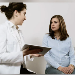 A white woman in a white lab coat talks to a young white woman in a blue button down shirt.