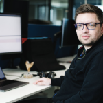 A young man with scruff, short brown hair and thick-framed glasses sits in front of a laptop and monitor in what appears to be an office setting. He wears a black fleece jacket.