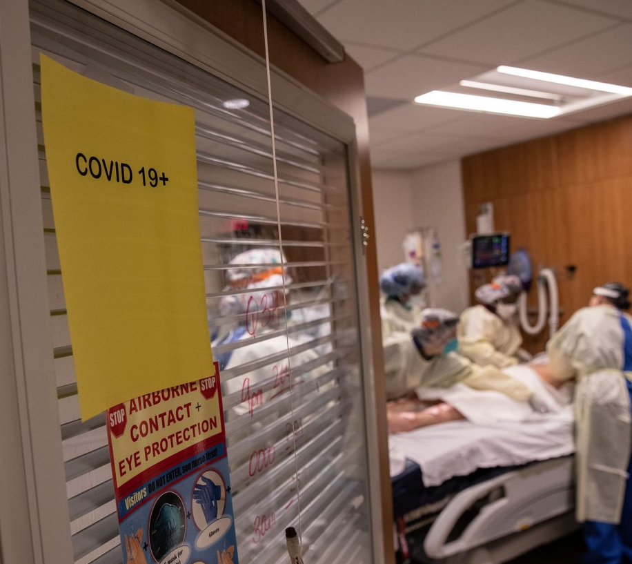 A hospital room with a COVID-19 sign posted on it. Healthcare workers gather around a patient inside.