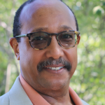 Ron Sibert headshot cropped to show just his face. Ron is a medium-skinned man with some grey in his mustache. He wears tortoiseshell framed glasses, a peach polo and light greige blazer.