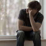 Adult male sitting in a window seat resting his head on his hand, looking contemplative.